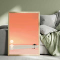 Framed artwork of a beach scene with a sunset over a body of water, placed on a sofa.